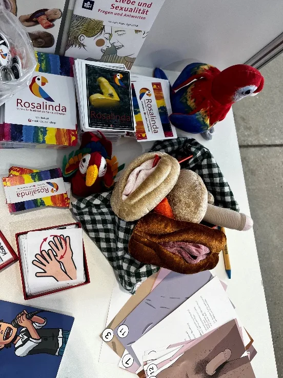 Tisch mit aufbereiteten Materialien von Rosalinda, Plüschgenitalien, Sticker, Kärtchen, Broschüren, Filzpapagei Rosalinda.