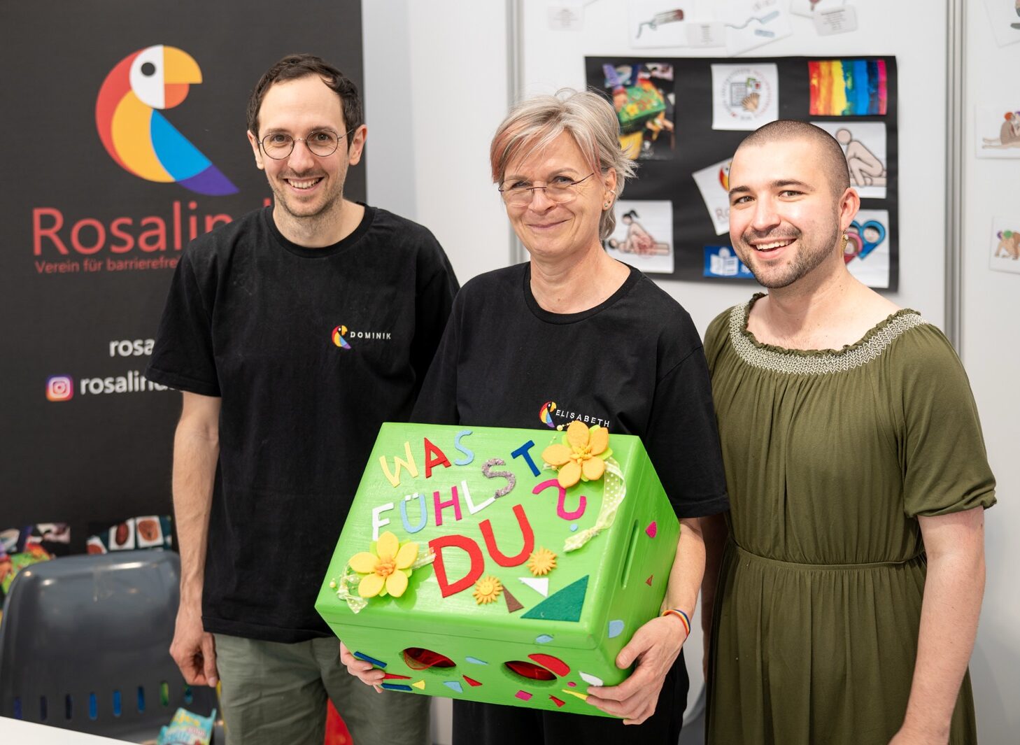 Mitglieder des Rosalinda Teams stehen lächelnd hinter einem Tisch mit verschiedenen Materialien und halten verschiedene Genitalmodelle sowie einen Plüschpapagei (Logo des Vereins) ins Bild. Hinter Ihnen befindet sich ein Rollup des Vereins sowie mehrere Plakate mit Metacom Icons zu den Themen Sex, Beziehung und Queer